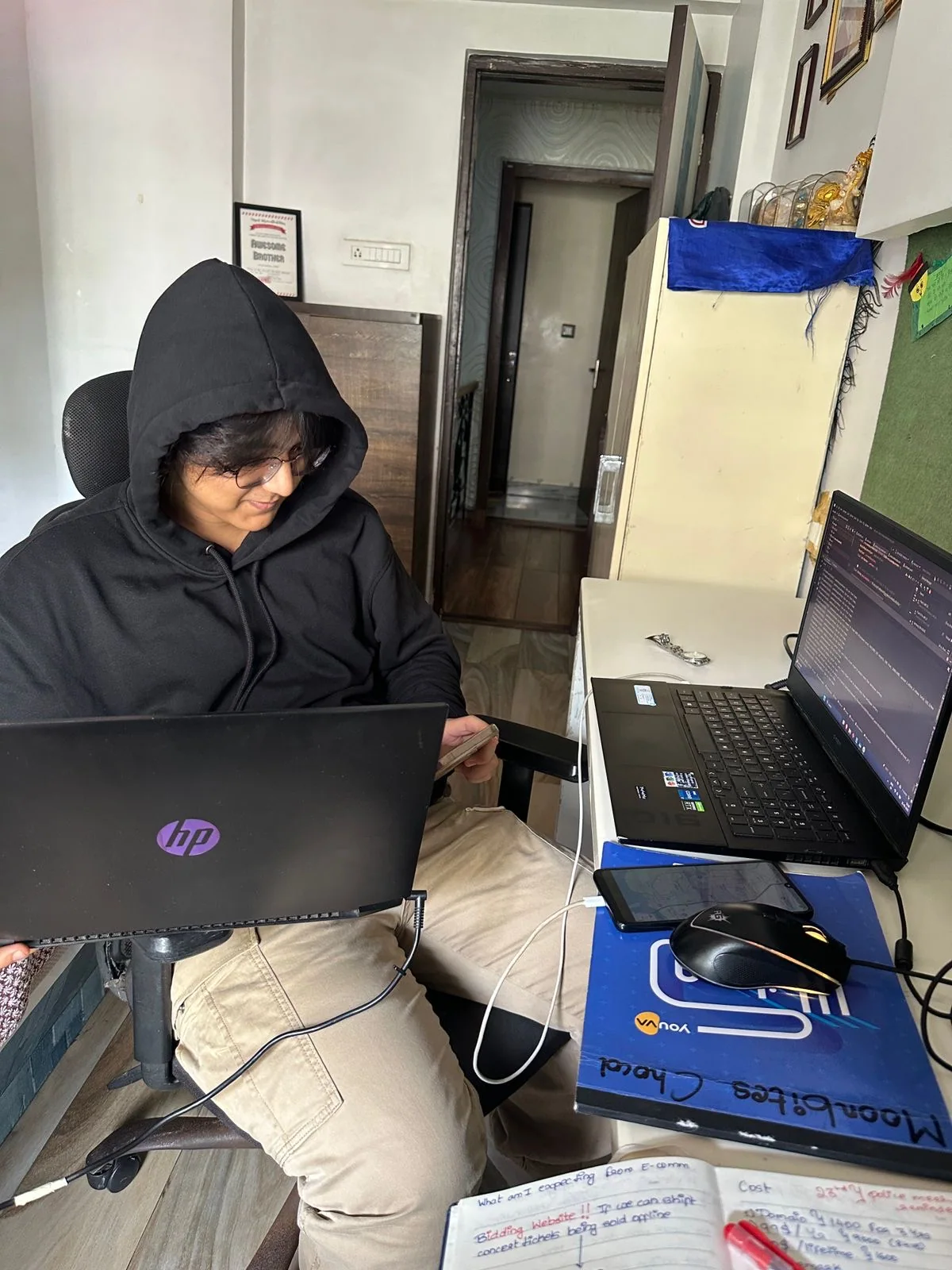 Aditya in a black hoodie working with two laptops at home